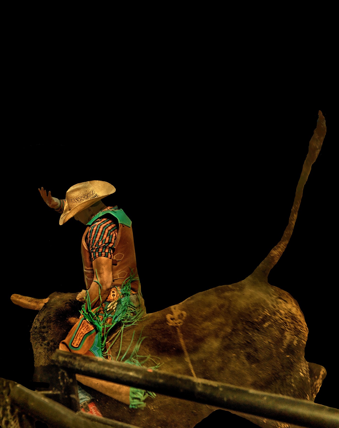 A bull rider wearing a cowboy hat competes atop a bull in a rodeo arena during a nighttime event with a dark background.