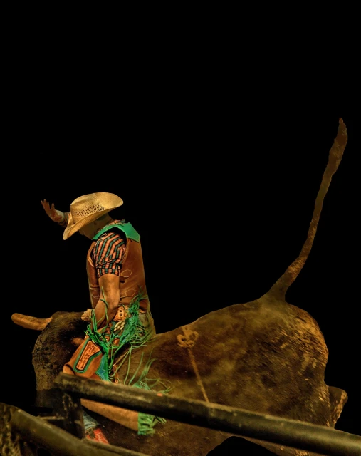 A bull rider wearing a cowboy hat competes atop a bull in a rodeo arena during a nighttime event with a dark background.