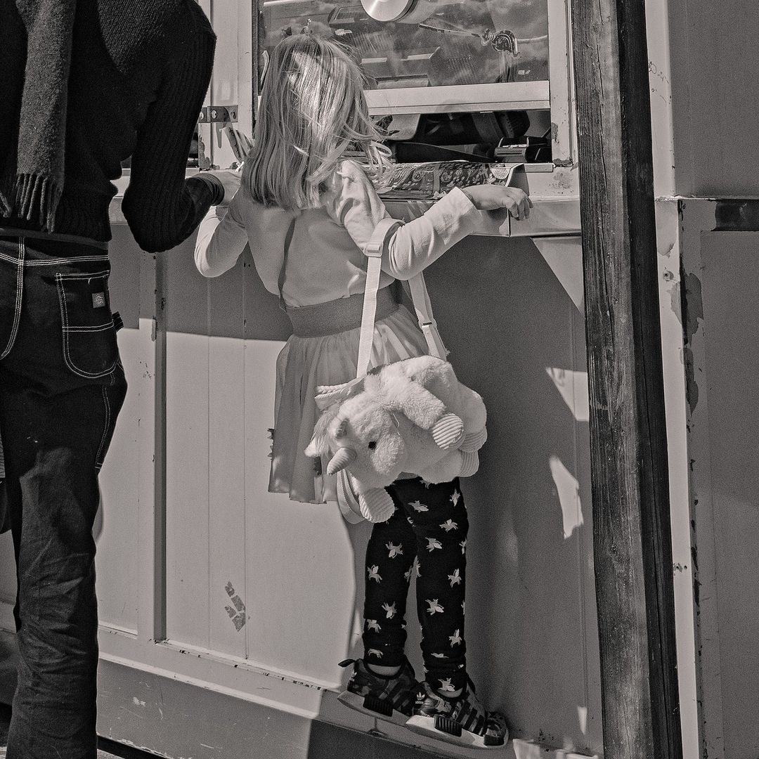 Black and white image of a child standing near a window with a stuffed animal purse, and an adult figure standing beside them.