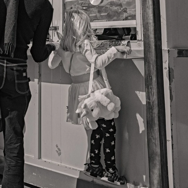 Black and white image of a child standing near a window with a stuffed animal purse, and an adult figure standing beside them.
