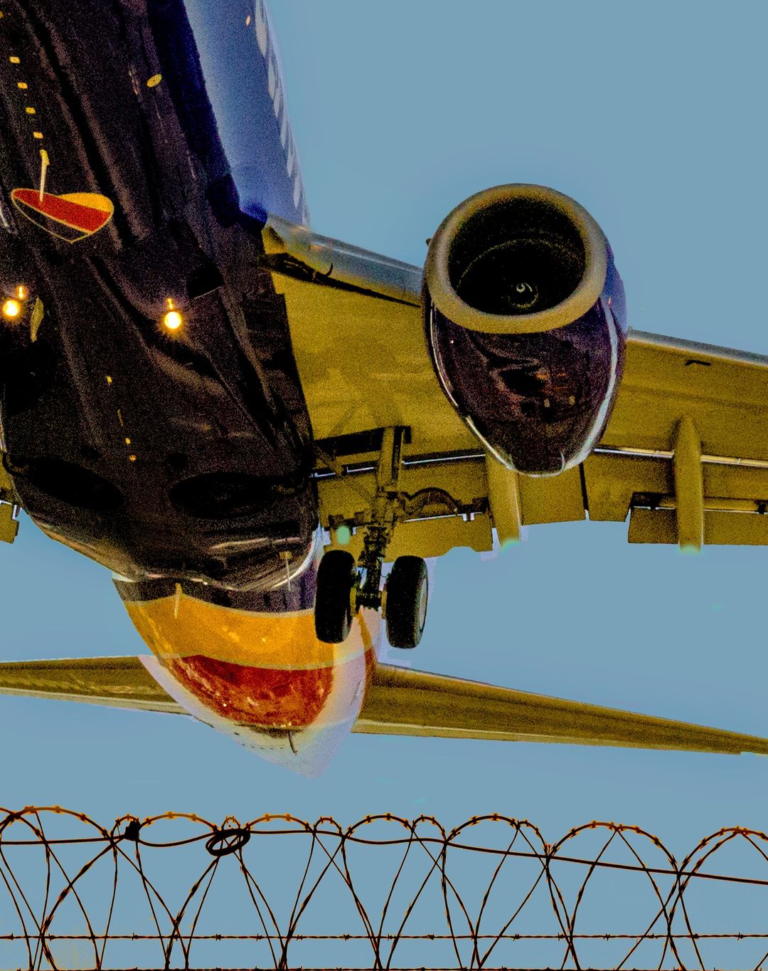 Low angle shot of a passenger airplane in flight, with a barbed wire fence in the foreground and a blue sky above.