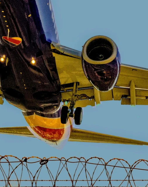 Low angle shot of a passenger airplane in flight, with a barbed wire fence in the foreground and a blue sky above.