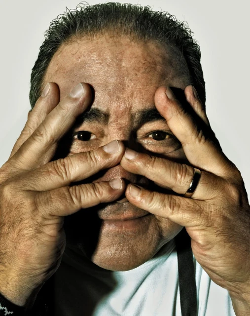 Headshot of an adult male looking through his fingers at the camera, wearing a white shirt and black suspenders in a studio.
