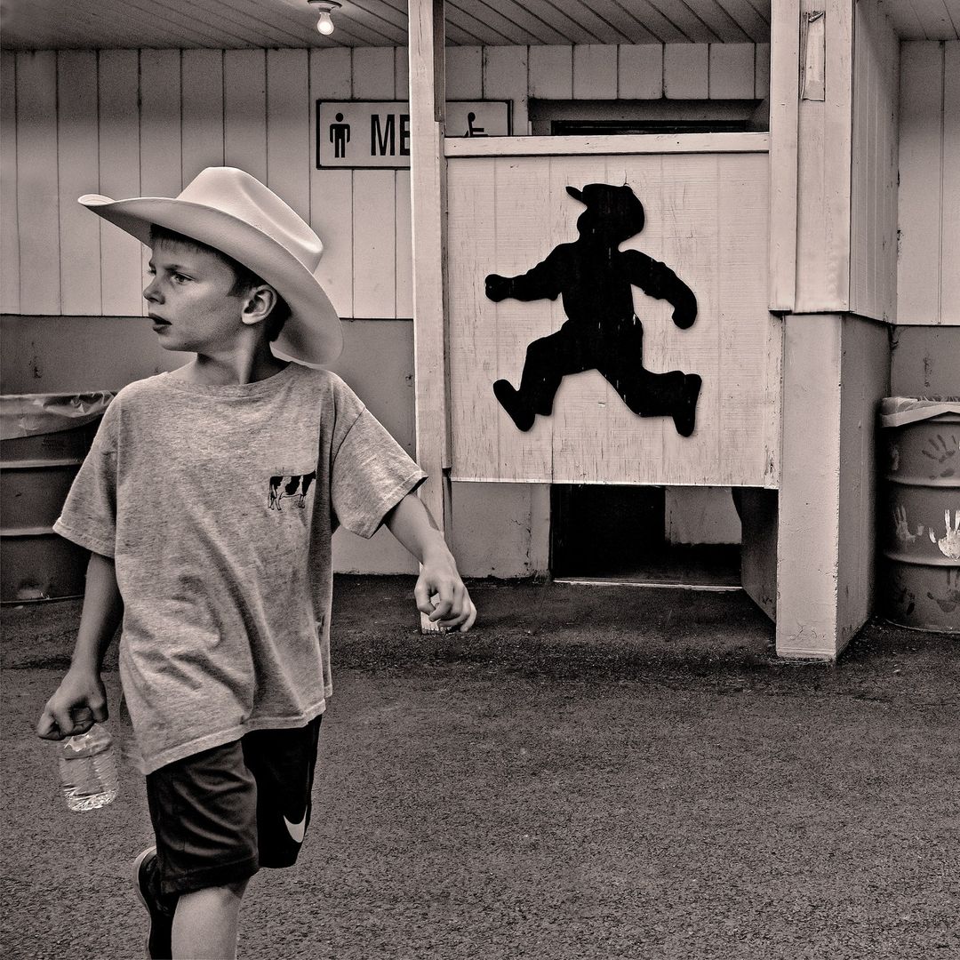 Black and white photo of a young boy with a cowboy hat and water bottle walking past a men's restroom with a running boy silhouette.