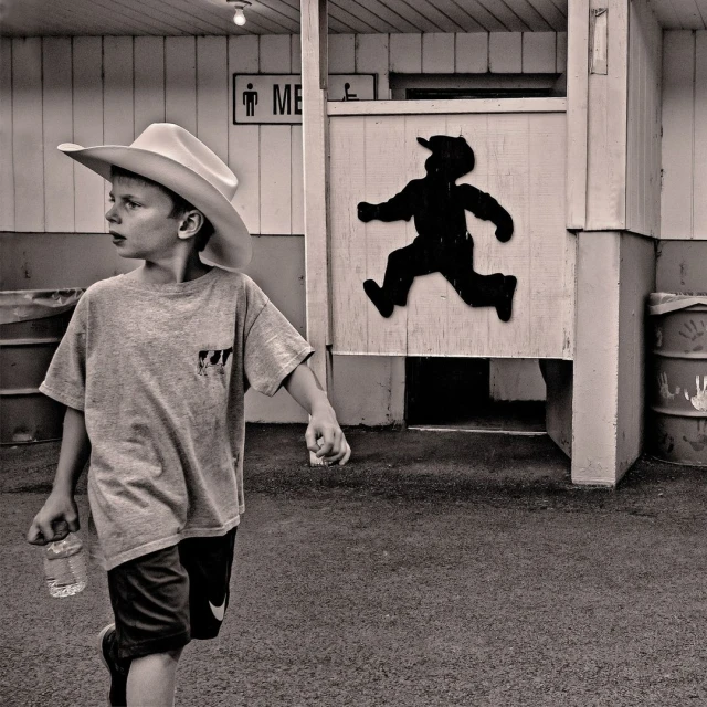 Black and white photo of a young boy with a cowboy hat and water bottle walking past a men's restroom with a running boy silhouette.