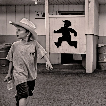 Black and white photo of a young boy with a cowboy hat and water bottle walking past a men's restroom with a running boy silhouette.