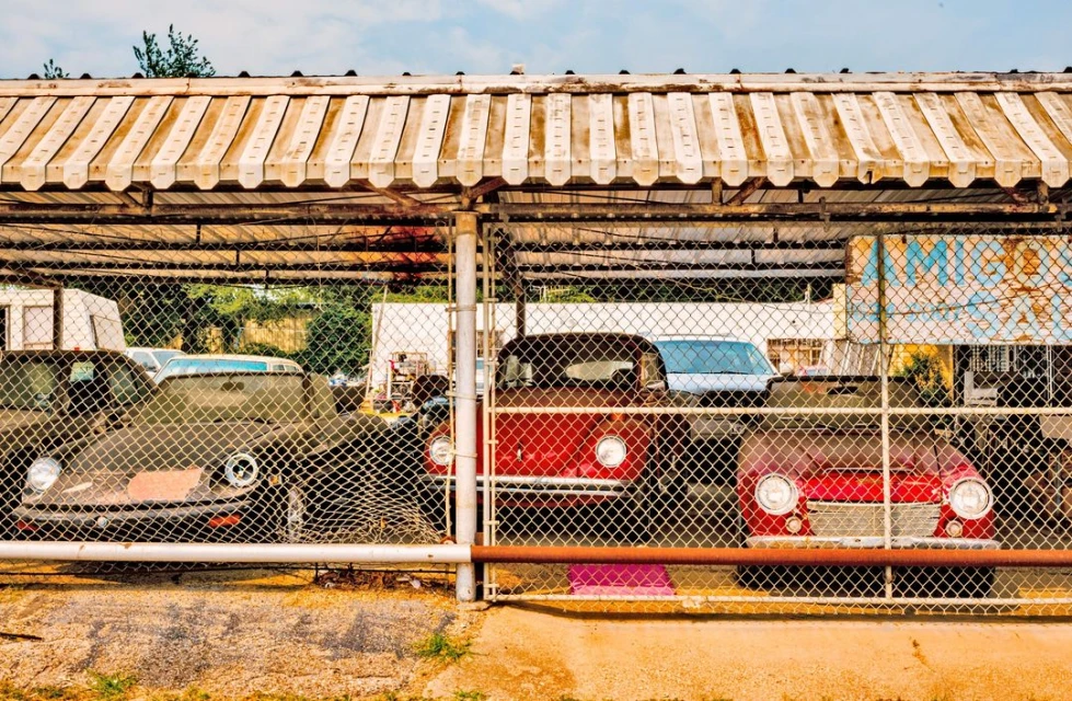 Vintage cars are parked behind a chain-link fence with a striped awning overhead, in a sunny outdoor setting, suggesting auto restoration or storage.
