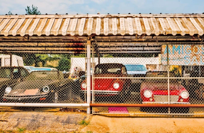 Vintage cars are parked behind a chain-link fence with a striped awning overhead, in a sunny outdoor setting, suggesting auto restoration or storage.