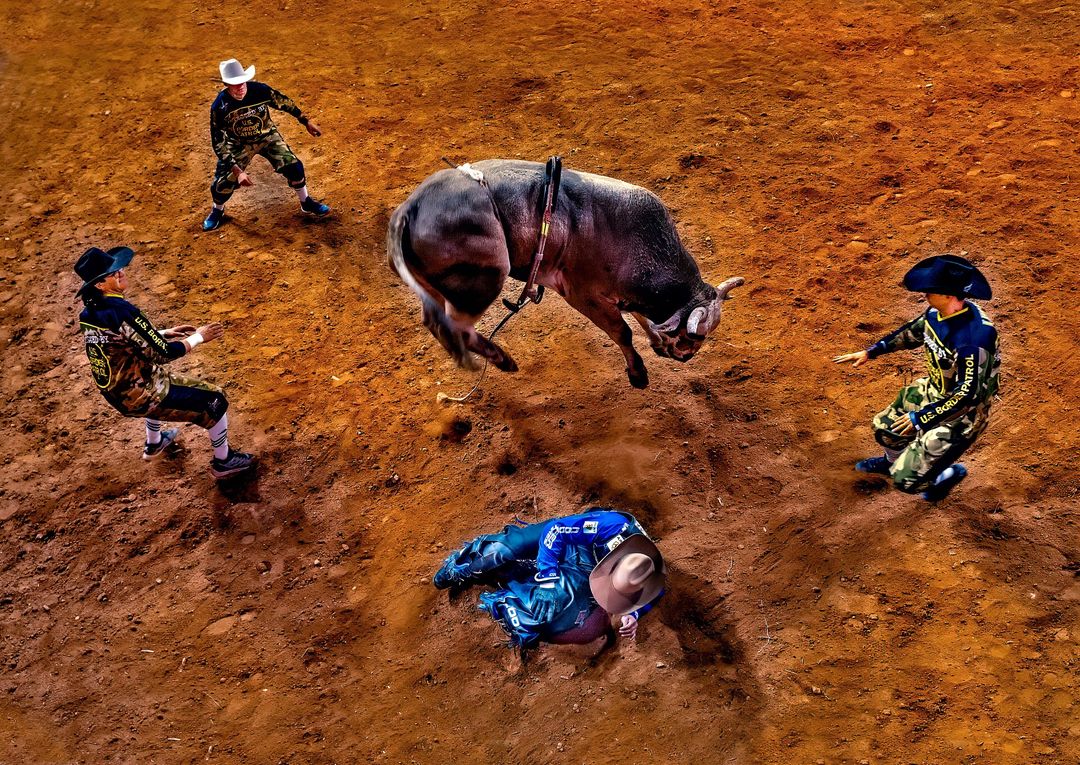 Dramatic overhead shot of a bull jumping in a rodeo arena with cowboys nearby; brown dirt ground and energetic scene, capturing the raw power.