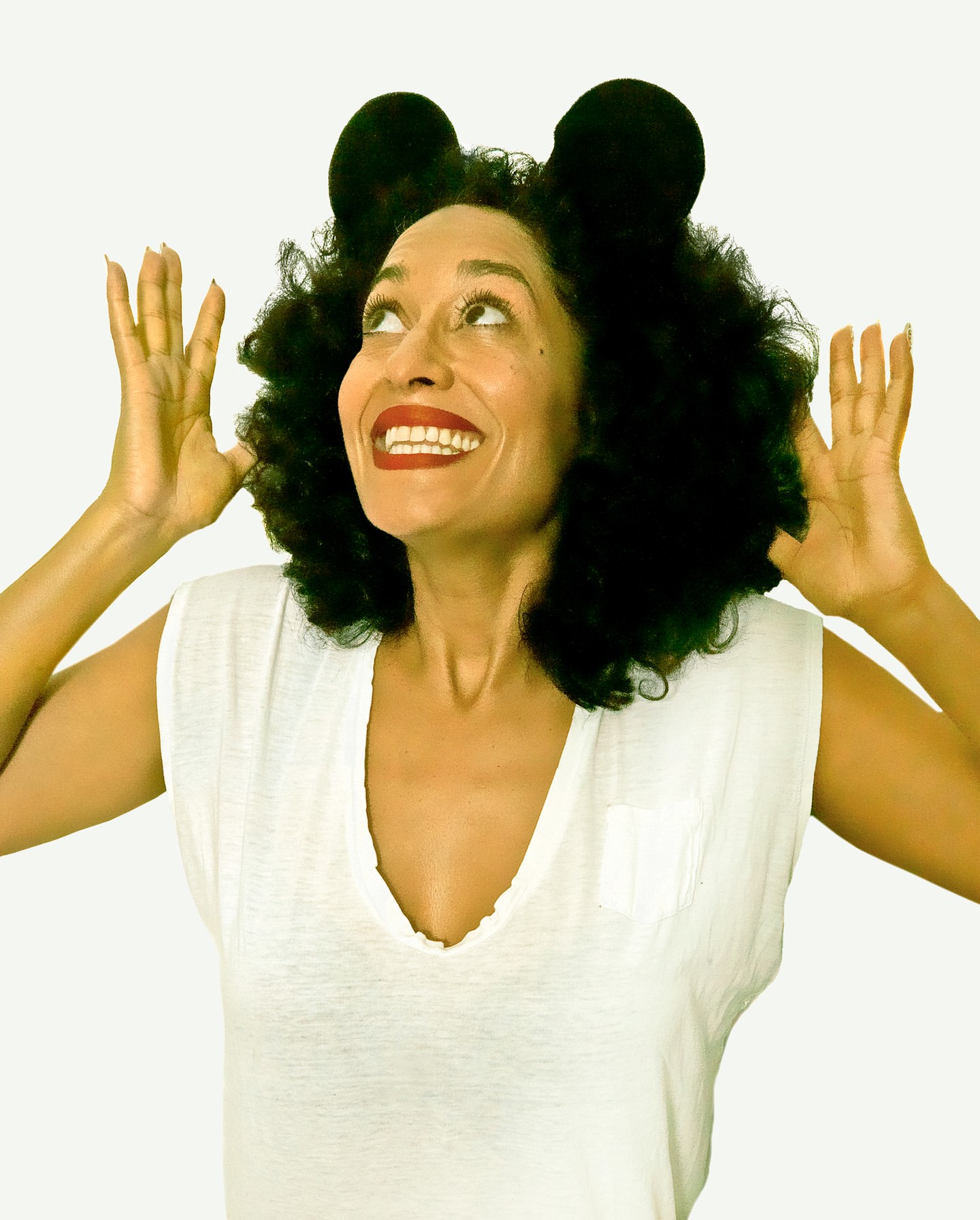 Smiling woman with afro wearing mouse ears, looking upwards with hands raised, posing against a bright white background. Studio shot.
