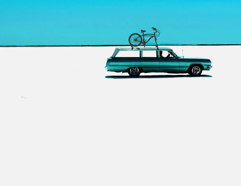 Aqua vintage station wagon with a bicycle mounted on top, parked in a flat, bright, minimalist landscape beneath a clear blue sky.