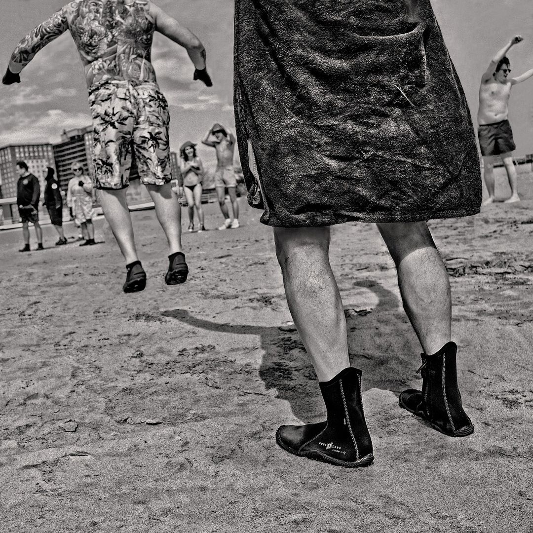 Monochrome image of people on a beach, focusing on a person in wetsuit boots and a towel wrapped around their waist, with others in swimwear.