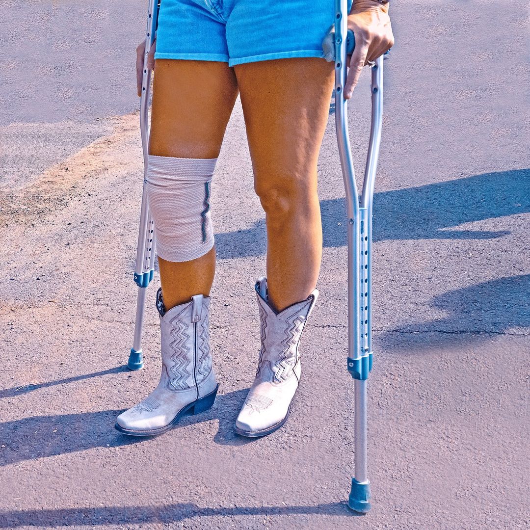 Woman standing with crutches and wearing cowboy boots and a knee brace. Outdoor shot. Legs and lower torso visible.