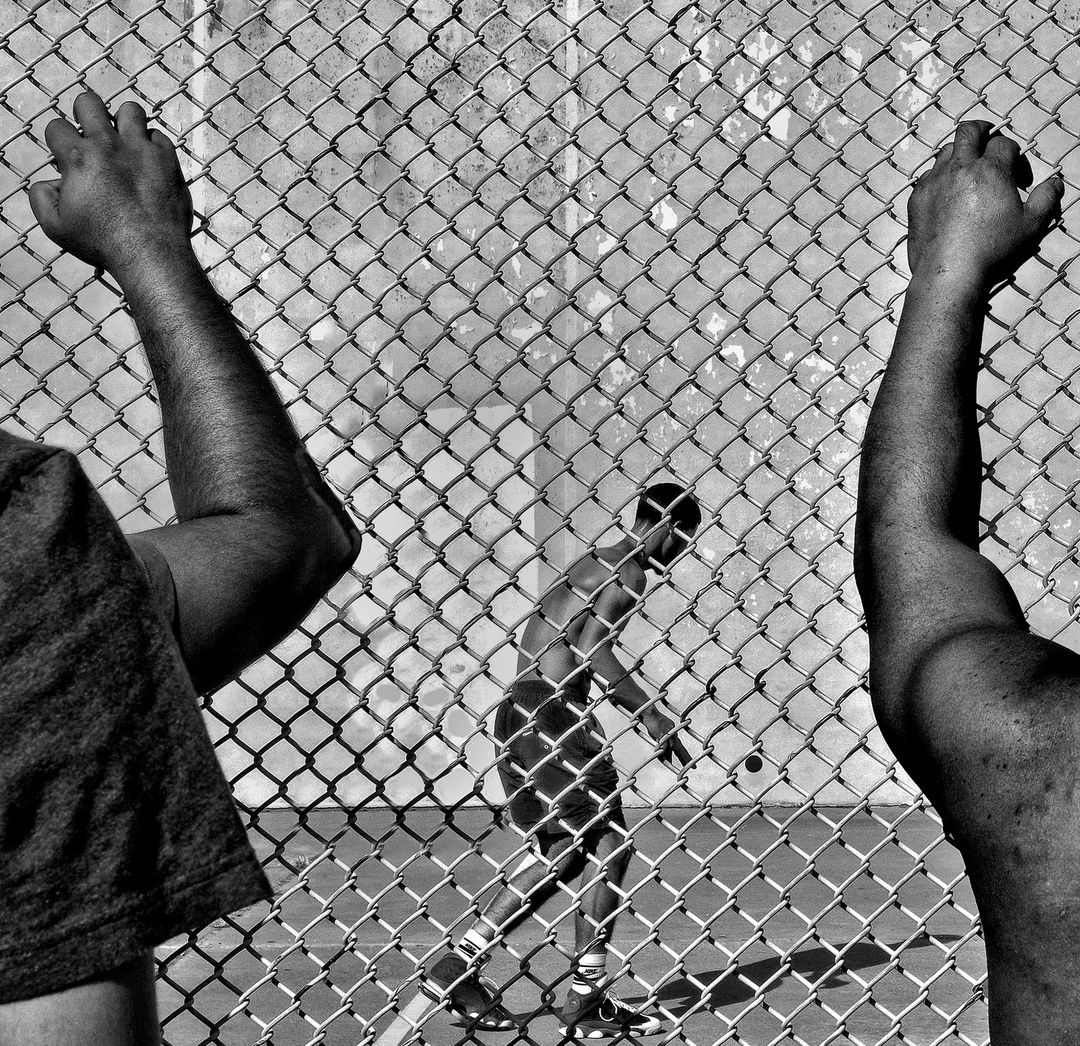 Monochrome shot of a man playing handball, framed by a chain-link fence with two partially visible figures on either side.