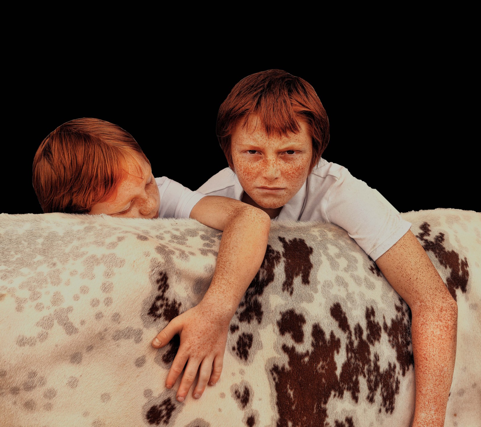 Two red-haired children with freckles resting on a brown and white spotted object against a black background in a close-up studio shot.