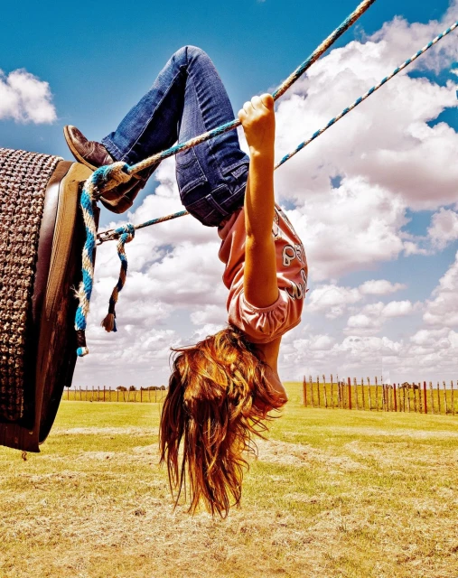 Girl hanging upside down on tire swing under a blue sky with clouds. She's wearing blue jeans and a pink shirt, enjoying an outdoor activity.