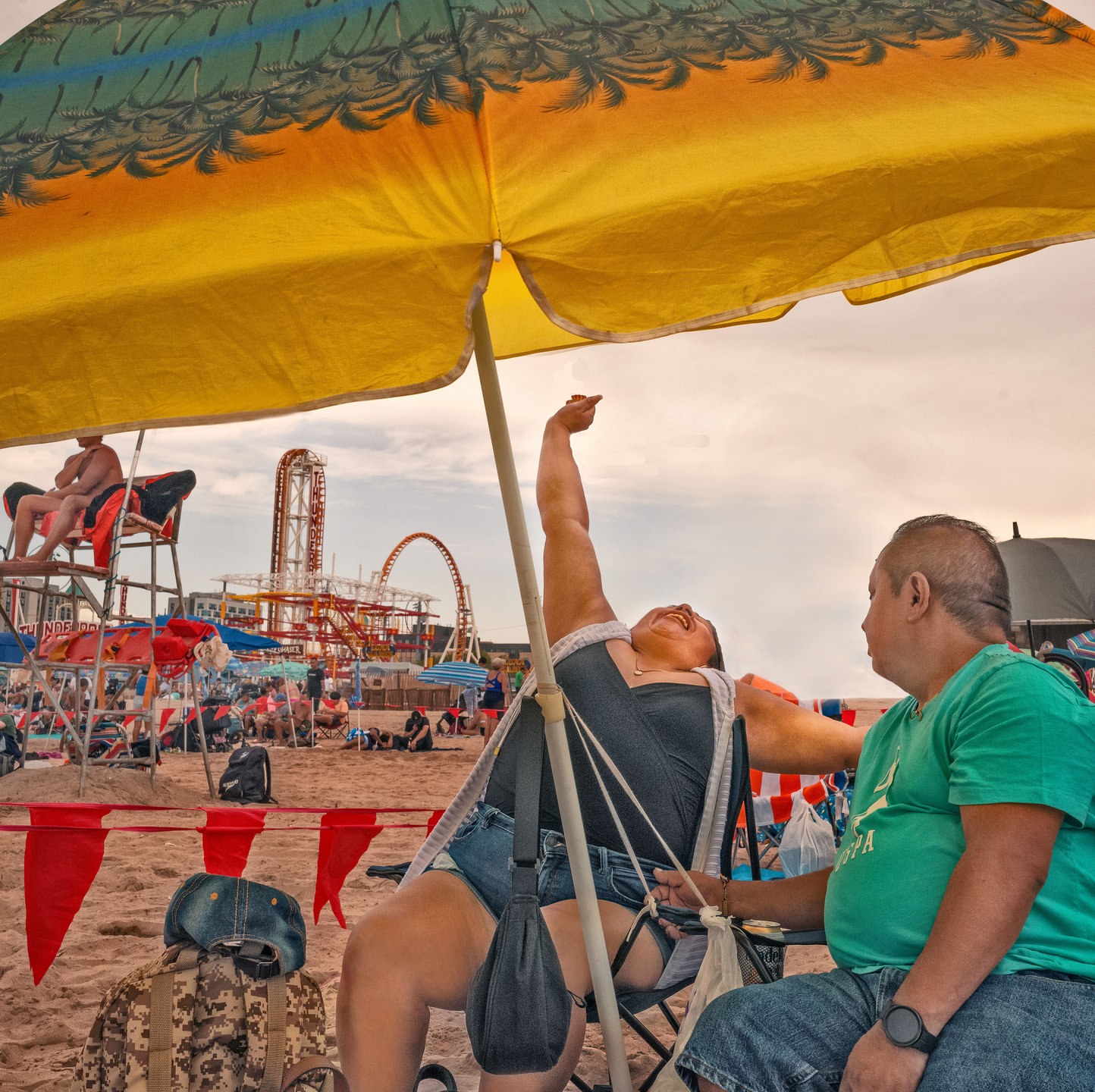 Two adults are relaxing on a sandy beach under a colorful umbrella, with a roller coaster visible in the background on a sunny day.