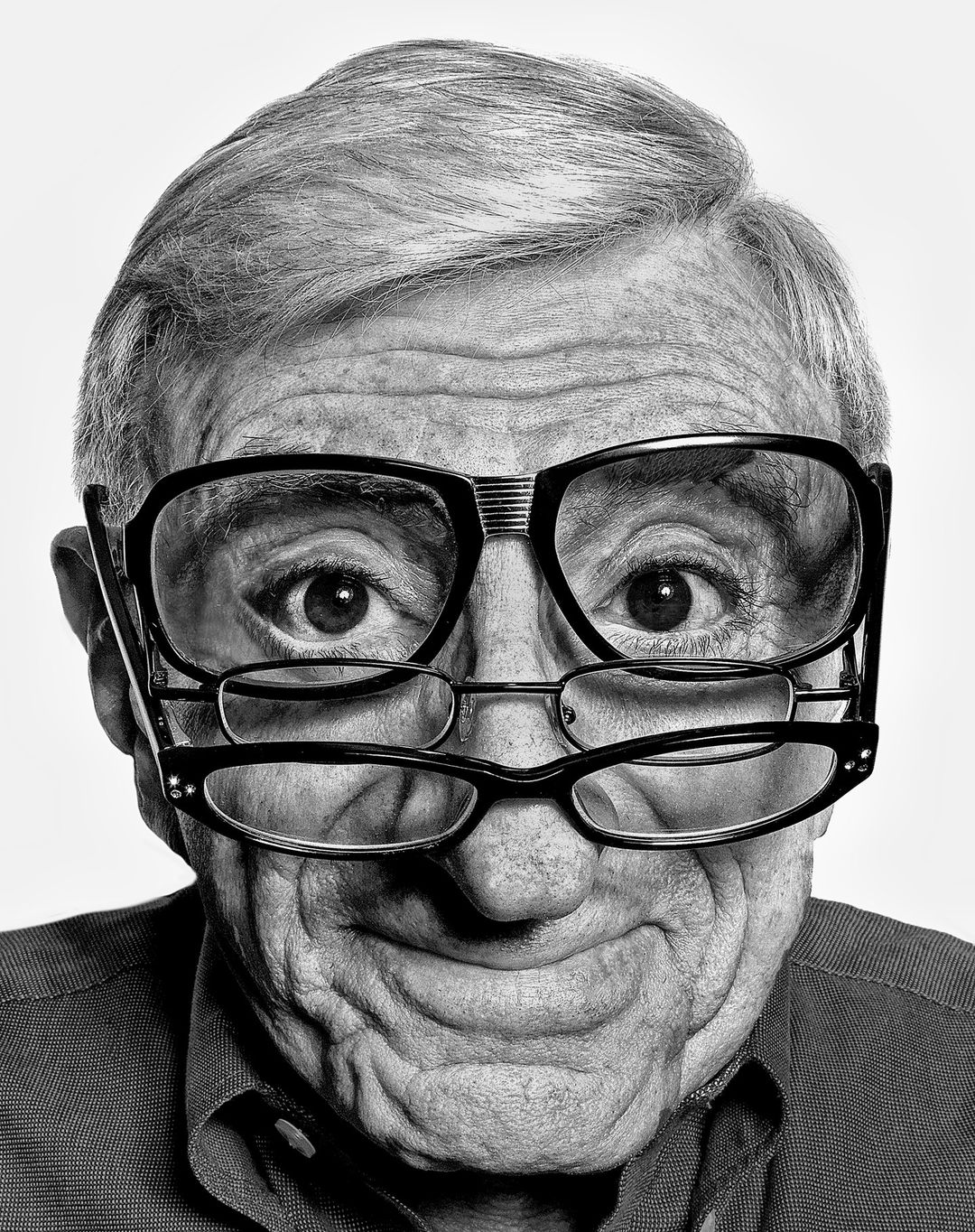 Black and white portrait of a senior man with two pairs of glasses, one resting on top of the other, against a plain white background.