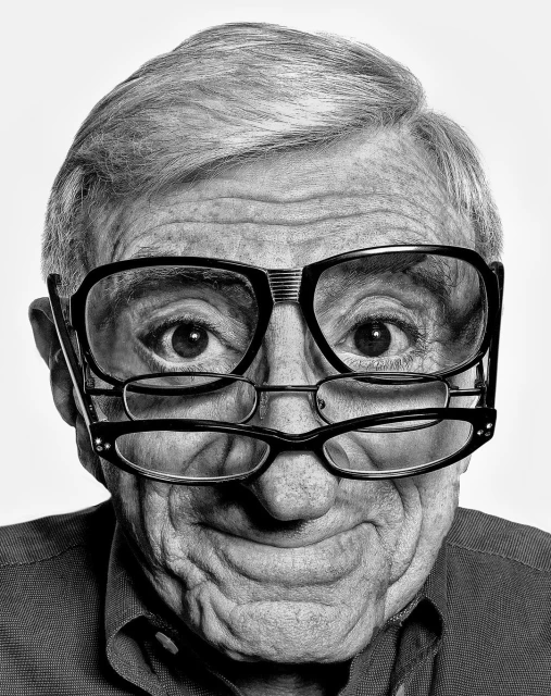 Black and white portrait of a senior man with two pairs of glasses, one resting on top of the other, against a plain white background.