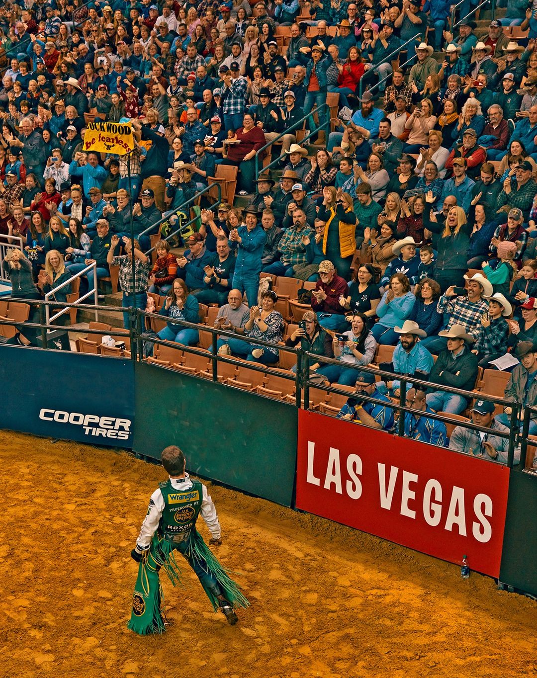 Bull rider in green chaps performs at a rodeo arena in Las Vegas with a crowd watching from the stands.