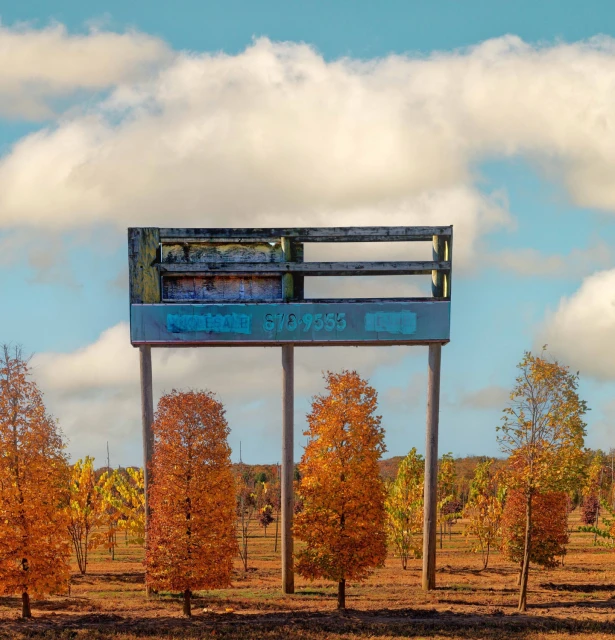 Weathered sign on wooden posts stands amidst a field of autumn-colored trees under a partly cloudy blue sky.