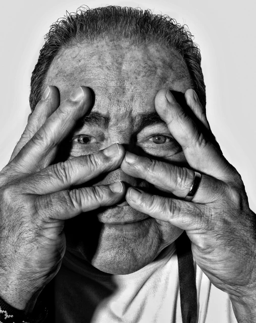 Monochrome portrait of an adult male with his hands framing his face, set against a bright background emphasizing the texture of the skin.
