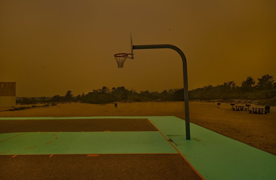 Outdoor basketball court with hoop and picnic tables under an orange sky, possibly from wildfire smoke, casting an eerie light.