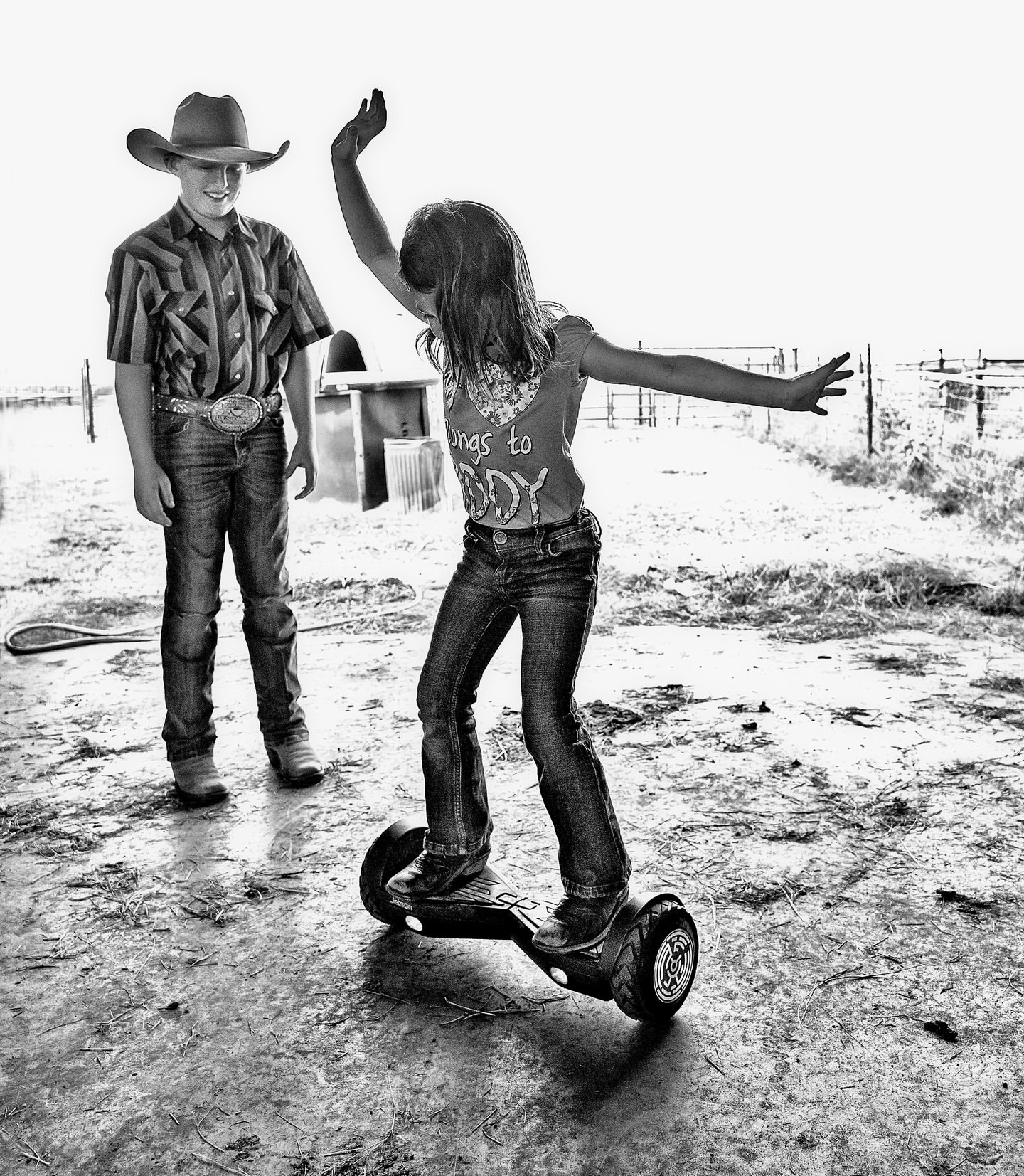 Black and white shot of a girl balancing on a hoverboard, with arms extended, while a boy in a cowboy hat watches in a rural setting.