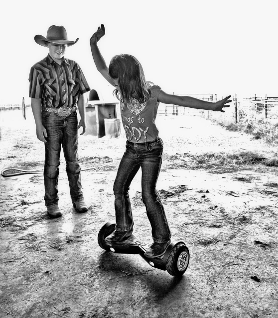 Black and white shot of a girl balancing on a hoverboard, with arms extended, while a boy in a cowboy hat watches in a rural setting.