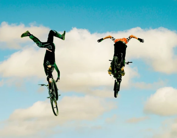 Two mountain bikers perform aerial stunts against a cloudy blue sky, showcasing skill and freedom in an outdoor sports scene.