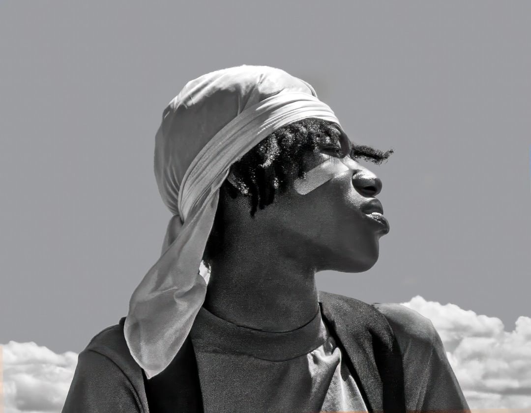 Black man wearing a silky durag and t-shirt against a cloudy sky background in New York by photographer Thom Gilbert.