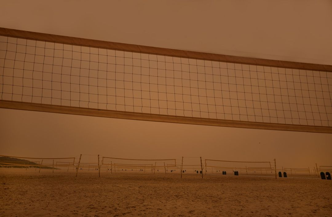 Multiple volleyball nets line a sandy beach under a hazy orange sky, creating a surreal and otherworldly atmosphere.
