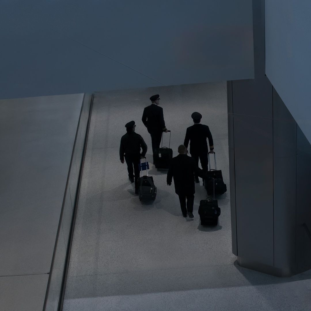 Four airline pilots in uniform walk with rolling luggage through an airport concourse in a low-lit, modern setting, viewed from above.