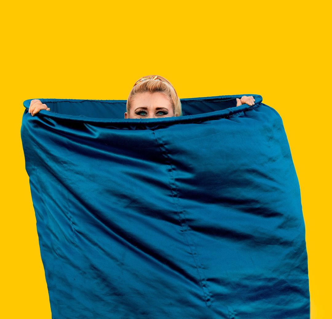 Woman peeking over the top of a shiny blue material against a bright yellow backdrop in a studio shot.