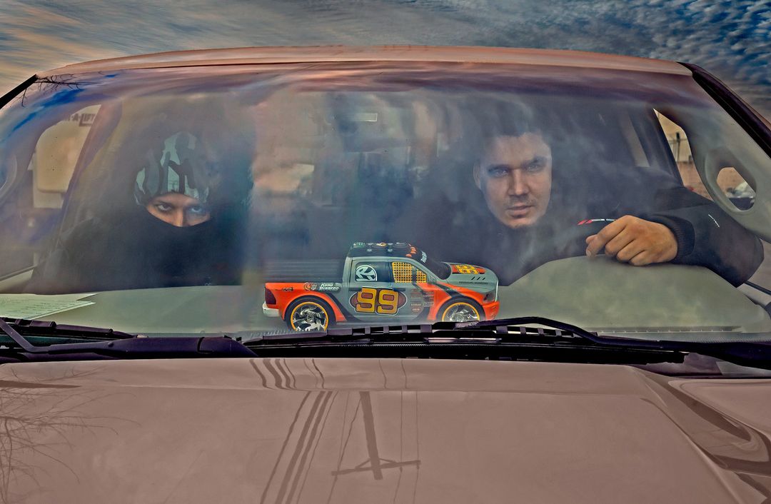 Two adults are visible through a car windshield, with a toy truck sitting on the dashboard. The sky is visible in the background.