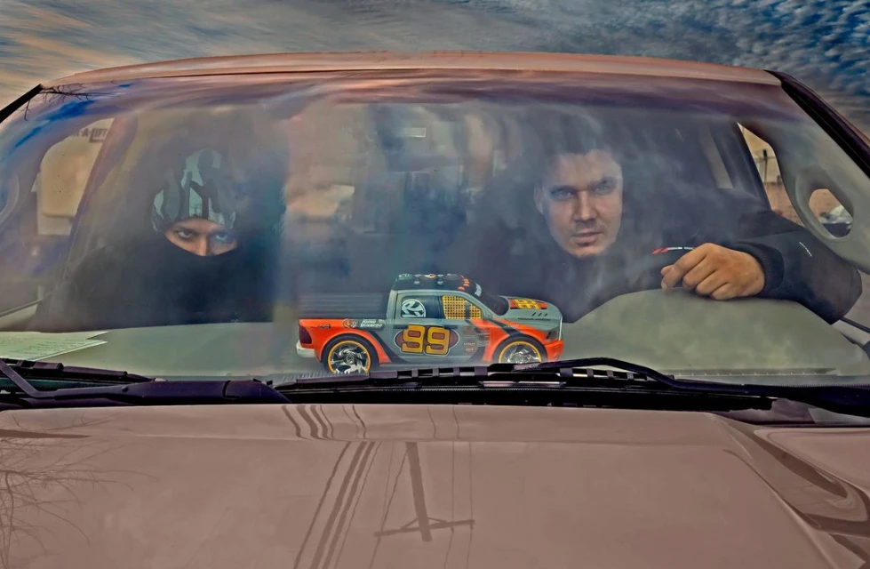 Two adults are visible through a car windshield, with a toy truck sitting on the dashboard. The sky is visible in the background.