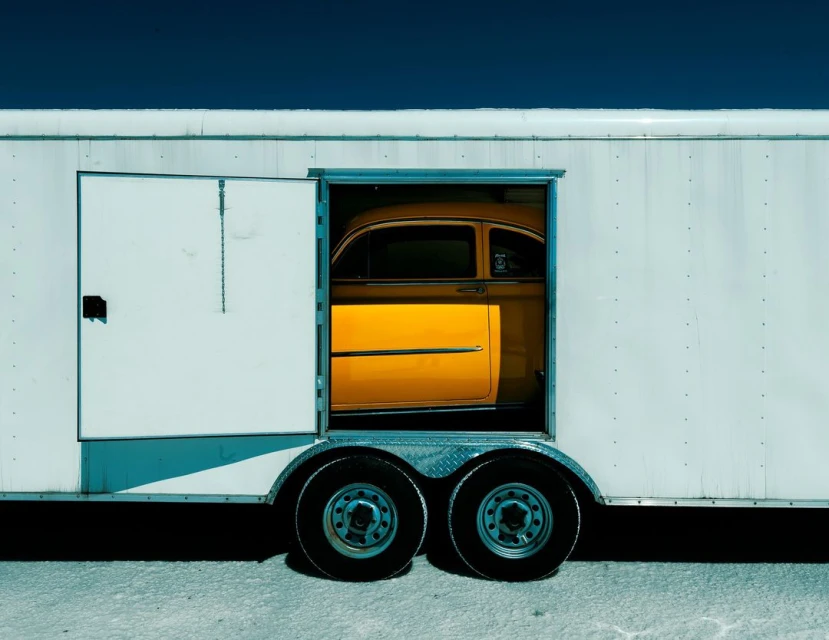 An old yellow car is partially visible inside a white trailer with an open door, set against a dark blue sky.