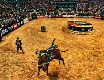 High-angle action shot of a cowboy riding a bucking bull in a dirt arena surrounded by stadium seating and brand banners.