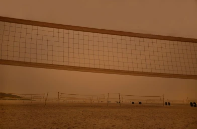 A volleyball net stretches across a hazy orange-tinted beach landscape under a thick monochromatic sky, photographed by Thom Gilbert.