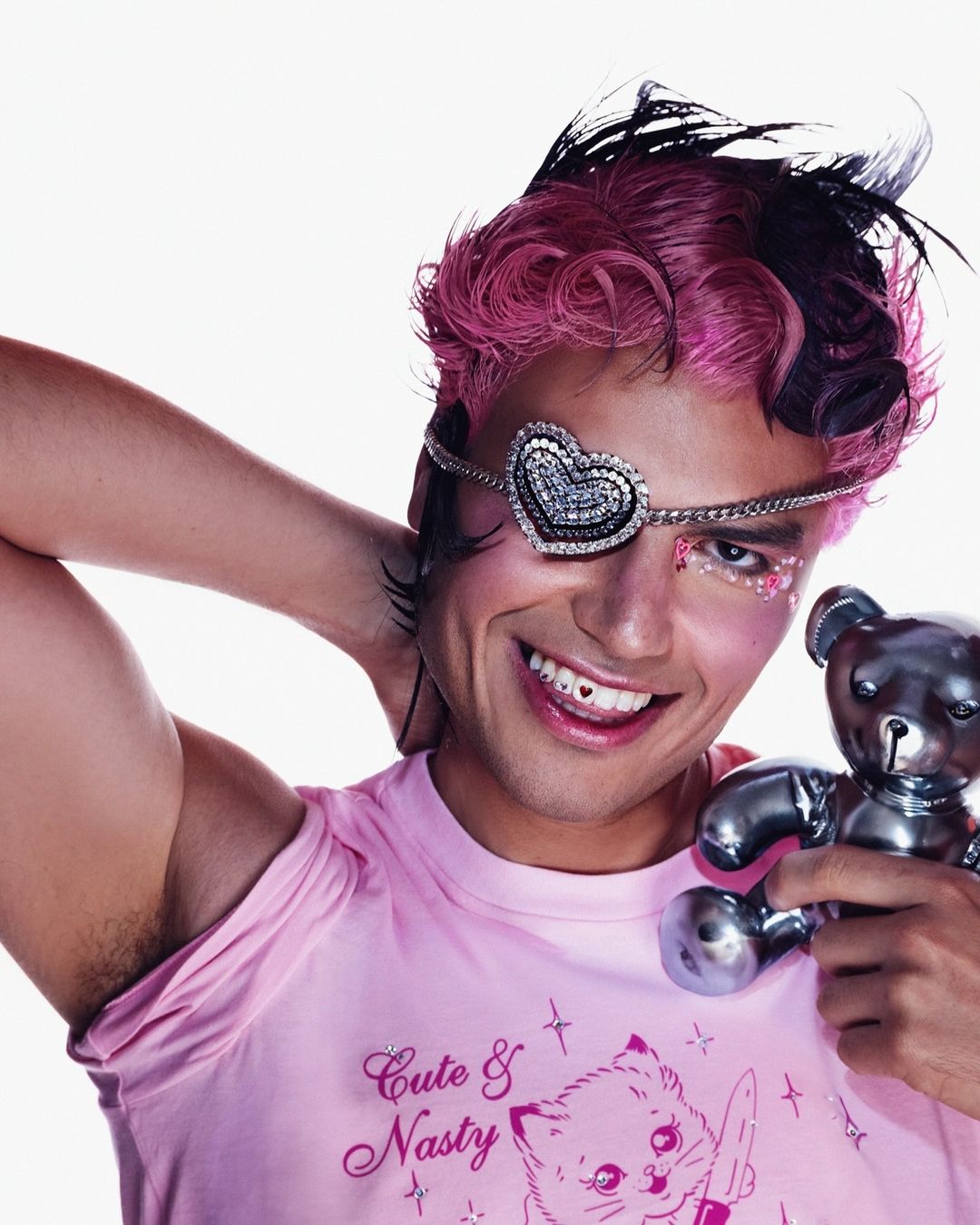 Smiling person with pink hair, eye makeup, and heart eyepatch, holding a metallic teddy bear against a plain white backdrop.