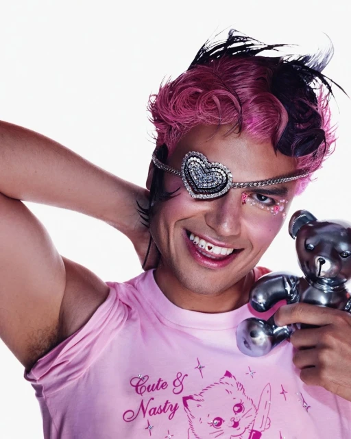 Smiling person with pink hair, eye makeup, and heart eyepatch, holding a metallic teddy bear against a plain white backdrop.