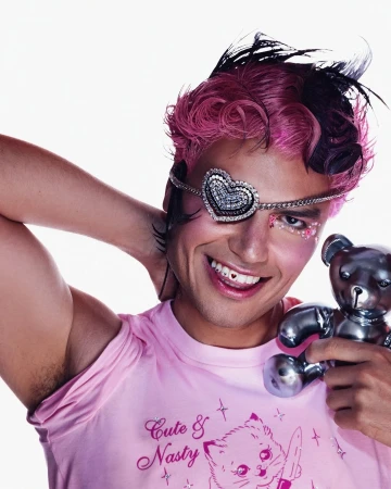 Smiling person with pink hair, eye makeup, and heart eyepatch, holding a metallic teddy bear against a plain white backdrop.