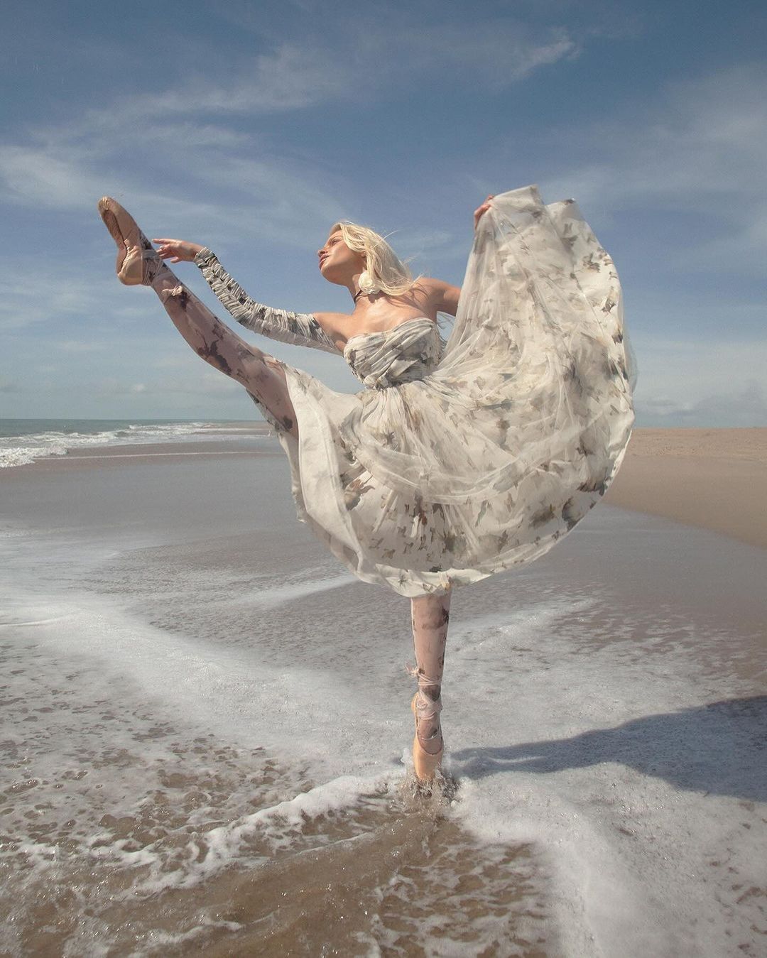 A woman in a flowing dress performs ballet in ocean shallows, stretching her leg high toward a partly cloudy blue sky.