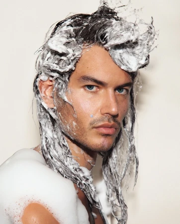 Portrait of a man with soapy foam in his dark hair and on his shoulder against a plain white background by Thom Kerr.
