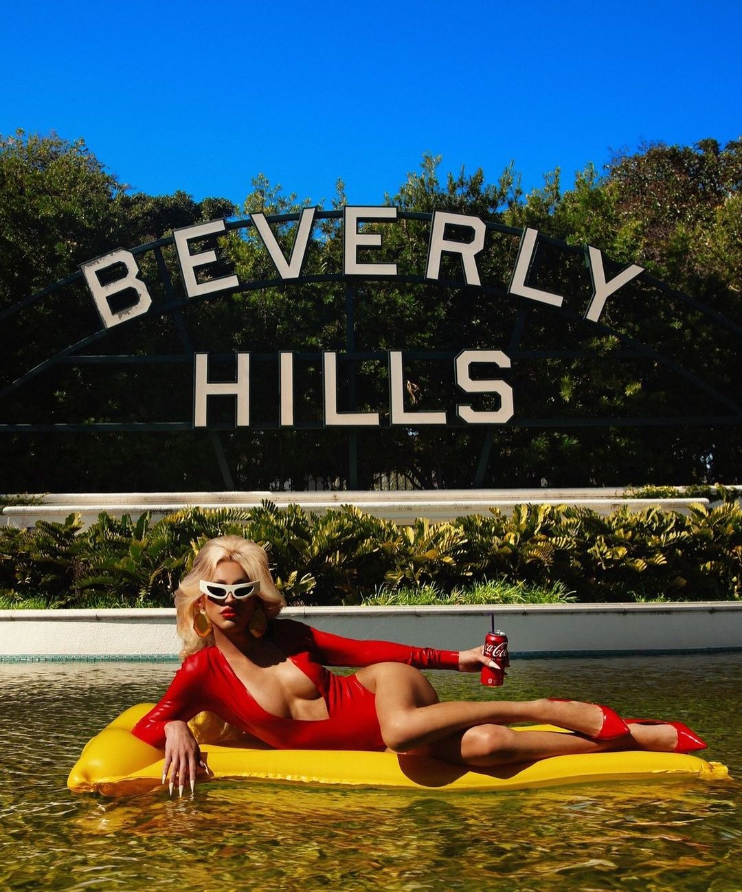 Woman in red swimsuit and sunglasses lounging on a yellow raft in a pool, holding a Coca-Cola, with the Beverly Hills sign in background.
