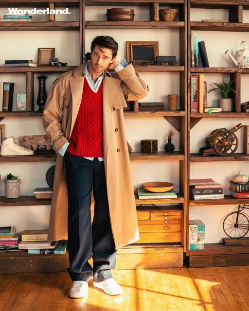 A stylish adult man poses in a trench coat with a red sweater vest against a wooden bookshelf in a studio setting. He is fashionable.