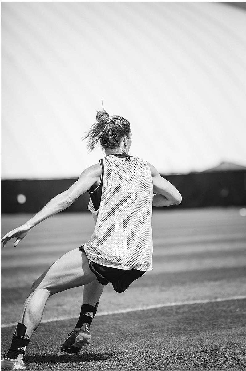 Monochrome image of a woman in athletic wear running on a grassy field with a large, white dome in the background on a sunny day.