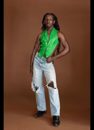 Black man wearing a vibrant green sleeveless top and light blue distressed jeans standing against a warm brown backdrop.