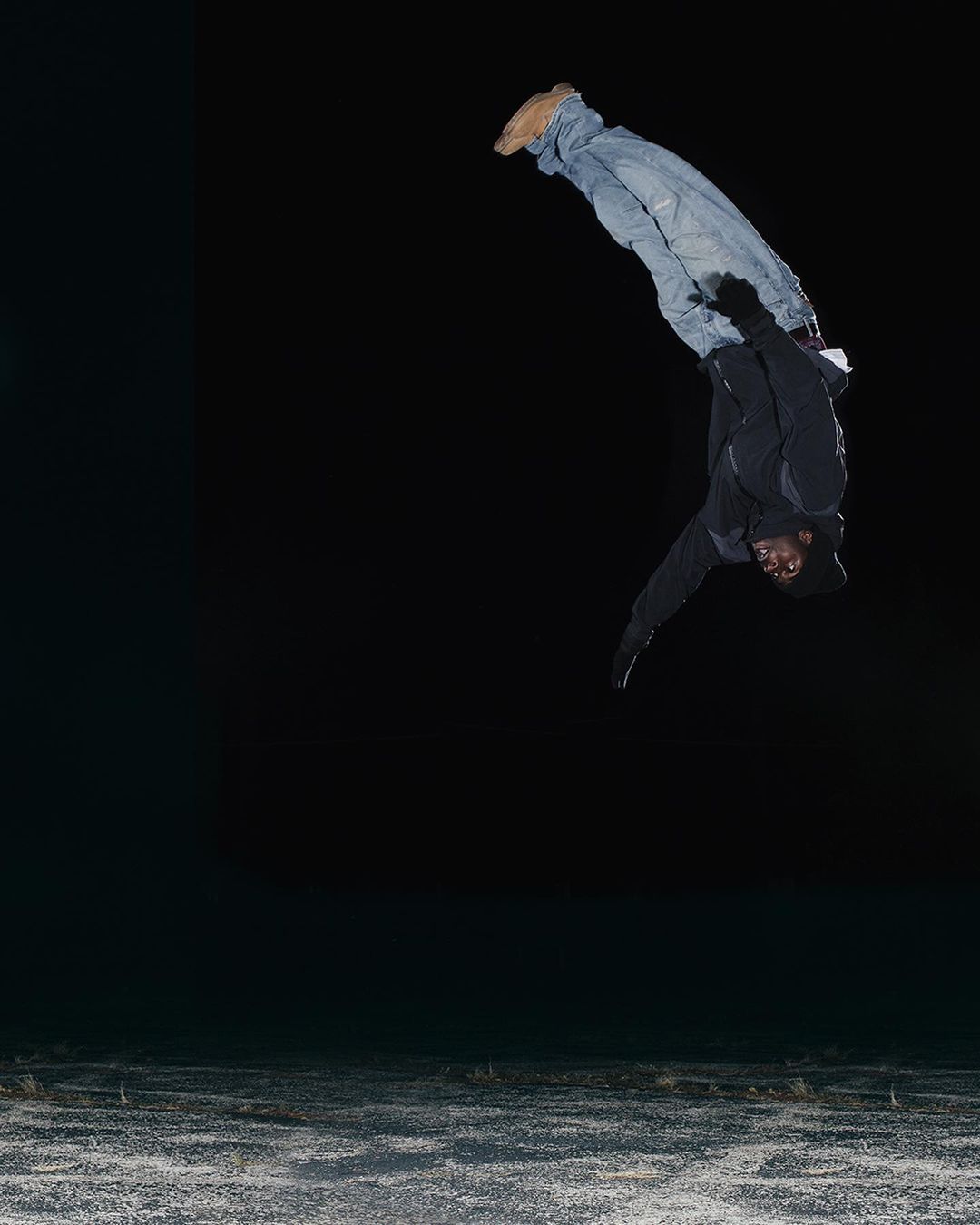 An adult male is captured mid-air, performing a backflip wearing a black jacket, blue jeans, and tan shoes against a dark backdrop.
