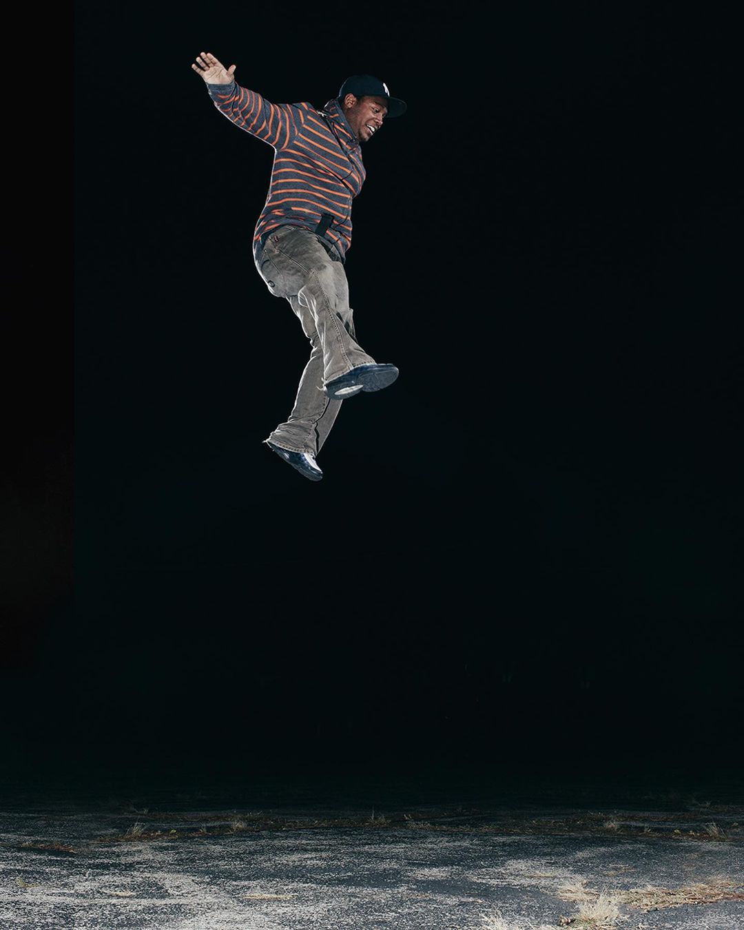 An adult male is captured mid-air, jumping with arms outstretched and a dynamic pose, against a stark black backdrop above a textured surface.
